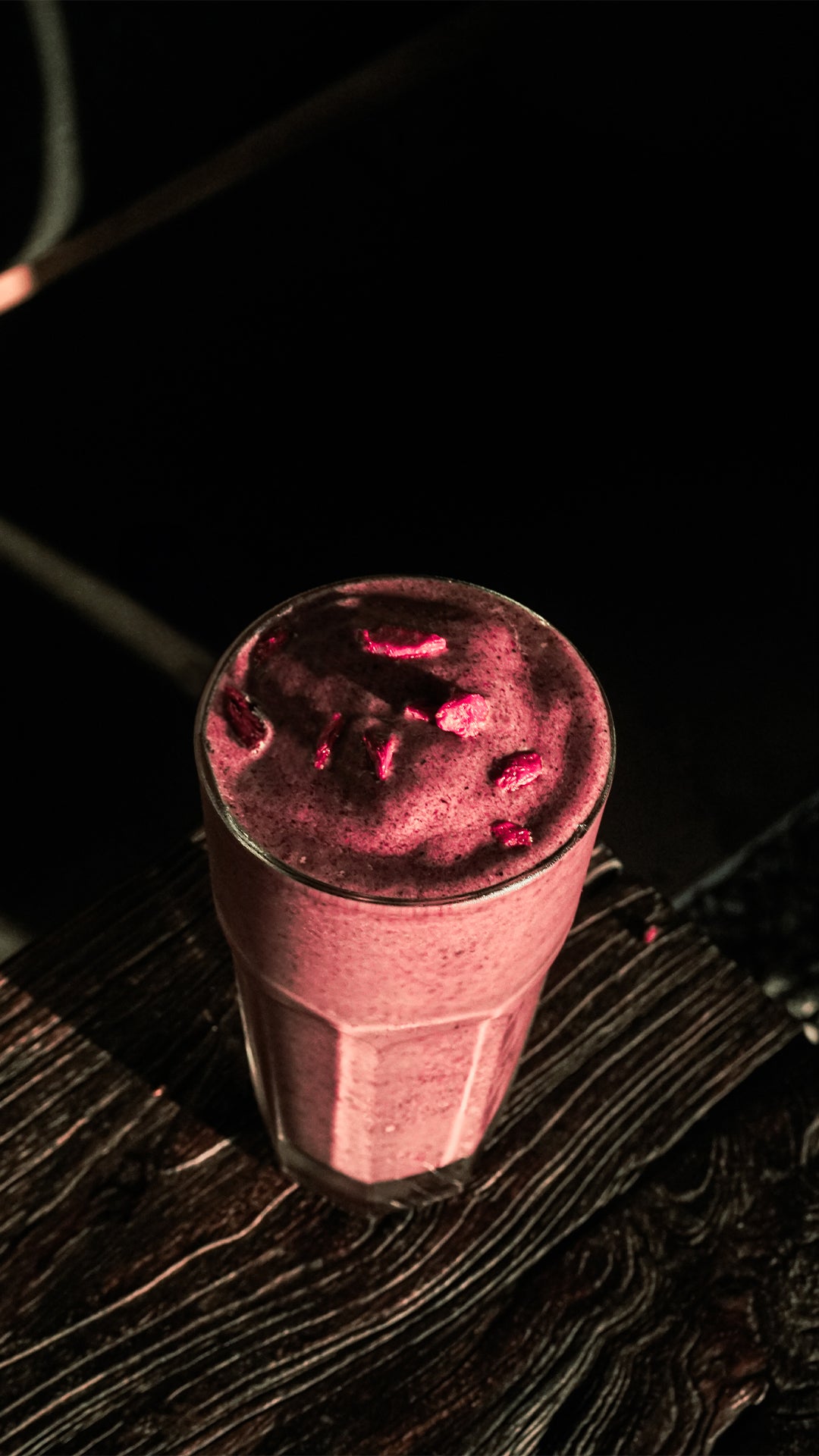 A vibrant berry smoothie in a tall glass, topped with bright red goji berries, placed on a rustic wooden table under dramatic lighting that highlights its rich texture and colour.