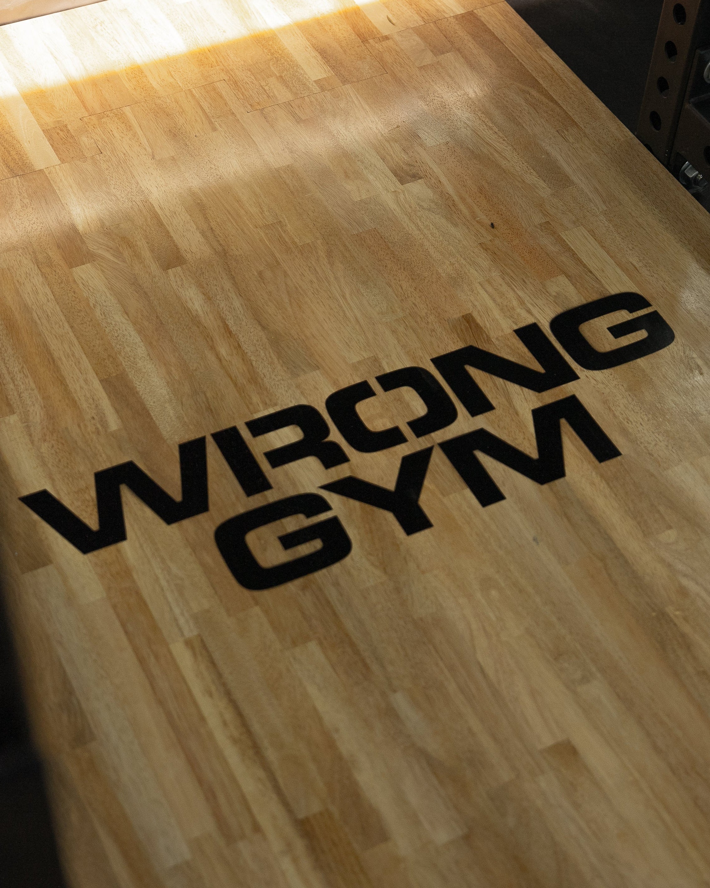 Close-up of a wooden gym platform with the bold black logo "WRONG GYM" printed across it, highlighted by soft natural light.