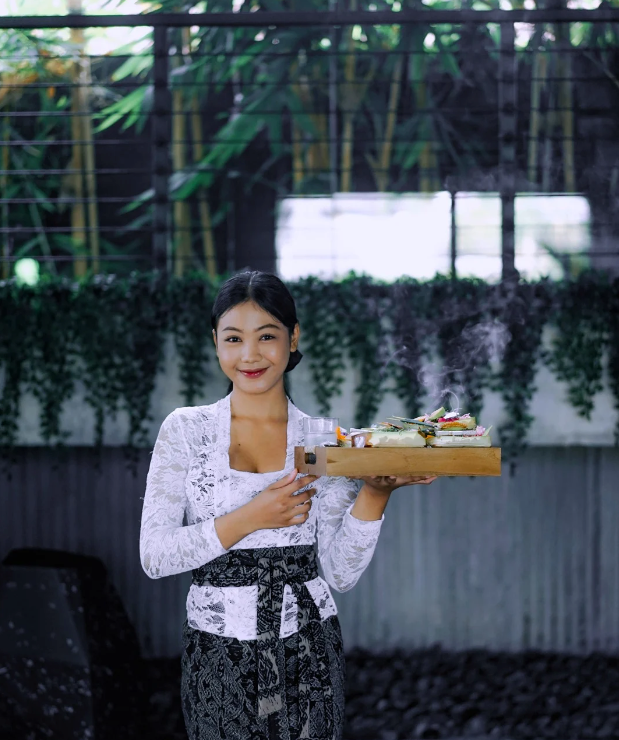 A smiling woman dressed in traditional Balinese attire holds a wooden tray with a steaming food offering. She stands in front of a wall adorned with lush green hanging plants and vertical bamboo structures, creating a serene and culturally rich backdrop.