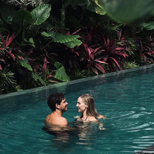 A couple smiling at each other while standing in a lush outdoor pool surrounded by tropical plants and vibrant greenery.