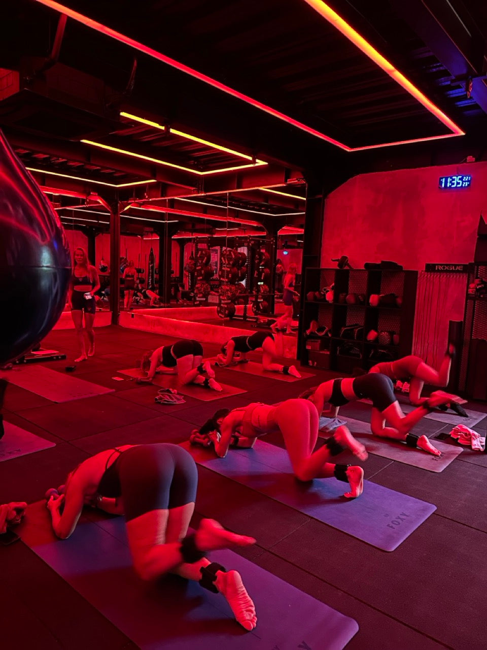 Group fitness class under red neon lighting with participants doing glute kickbacks on yoga mats, wearing ankle weights in a boutique gym setting.