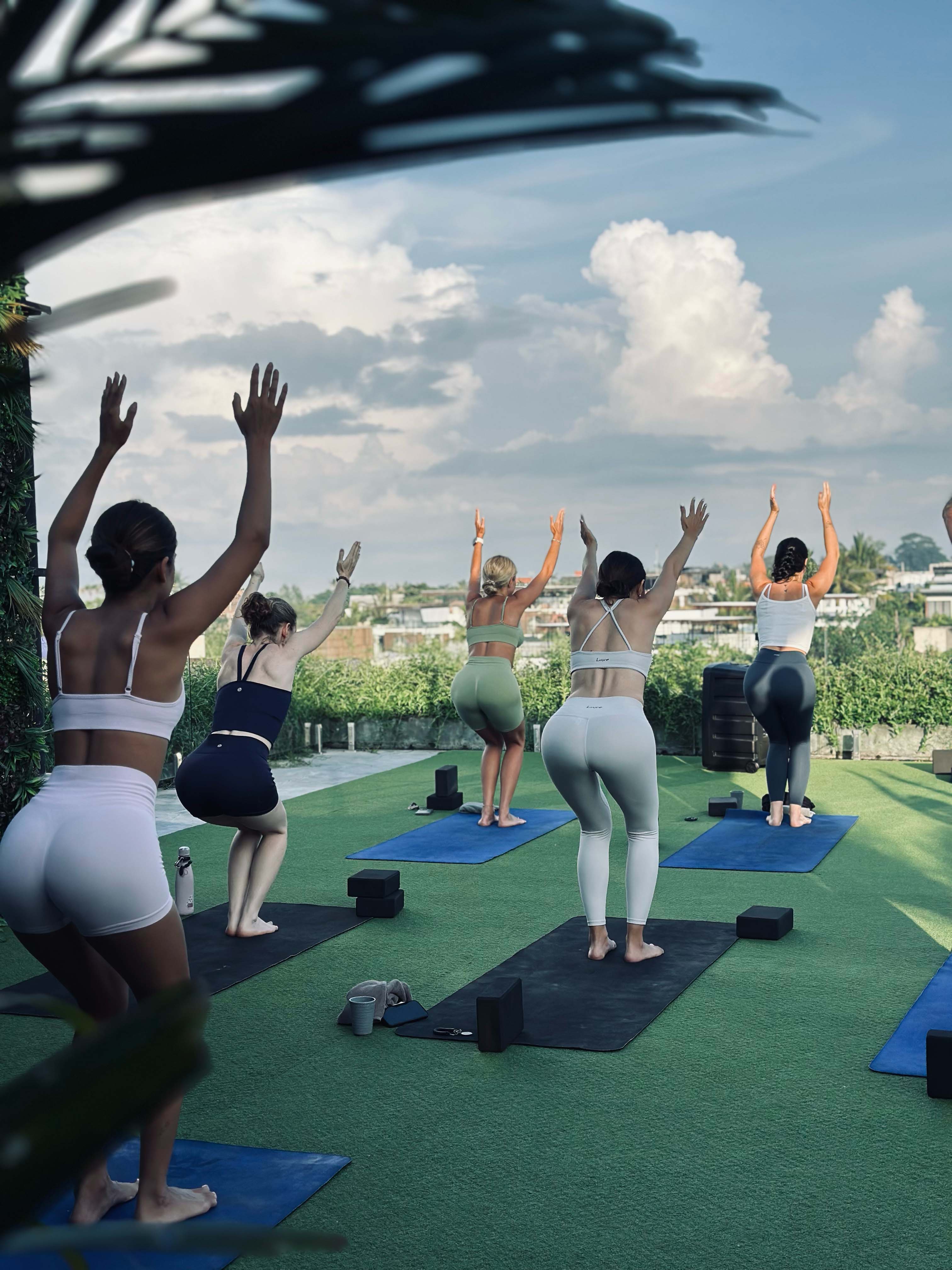 Group of women practicing yoga outdoors on a rooftop with blue mats and green artificial turf, facing a scenic skyline under a partly cloudy sky.