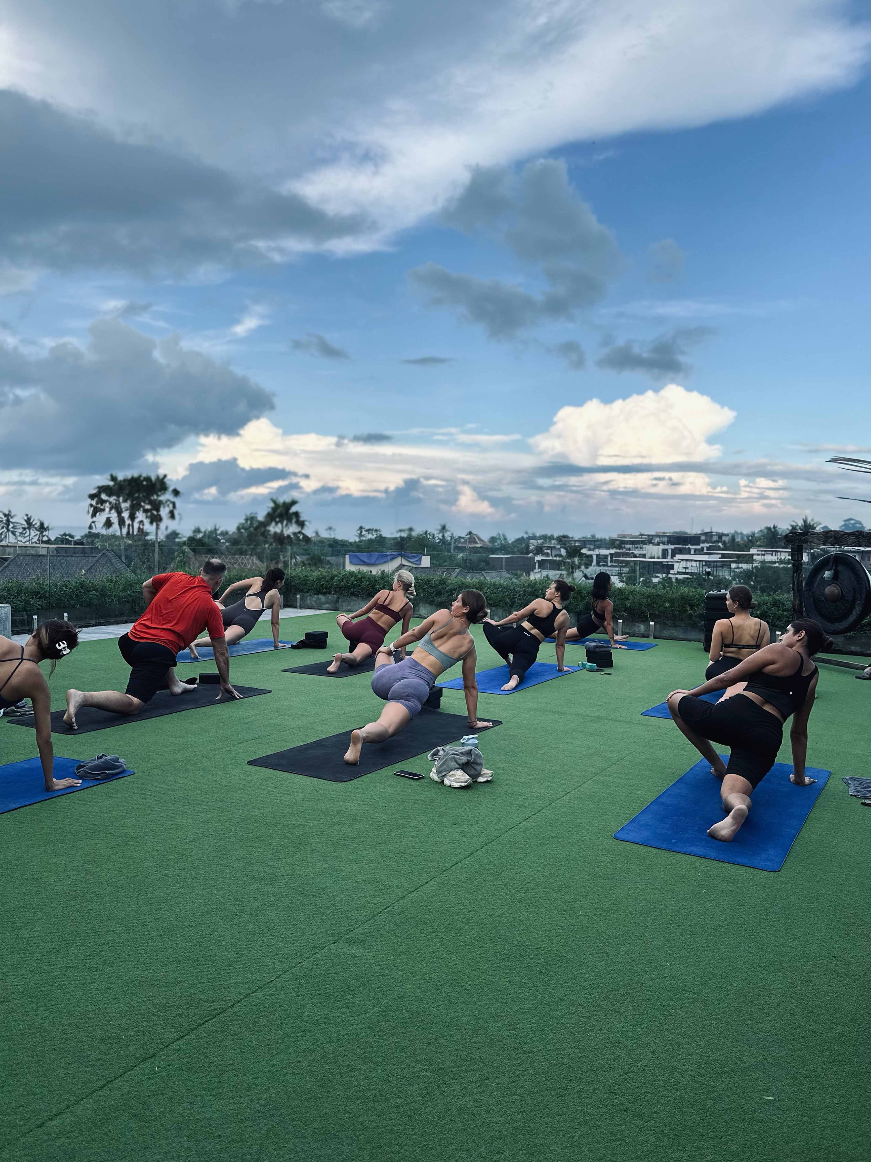 Group of people practising yoga on a rooftop with green turf and blue mats under a partly cloudy sky, surrounded by a scenic tropical landscape.