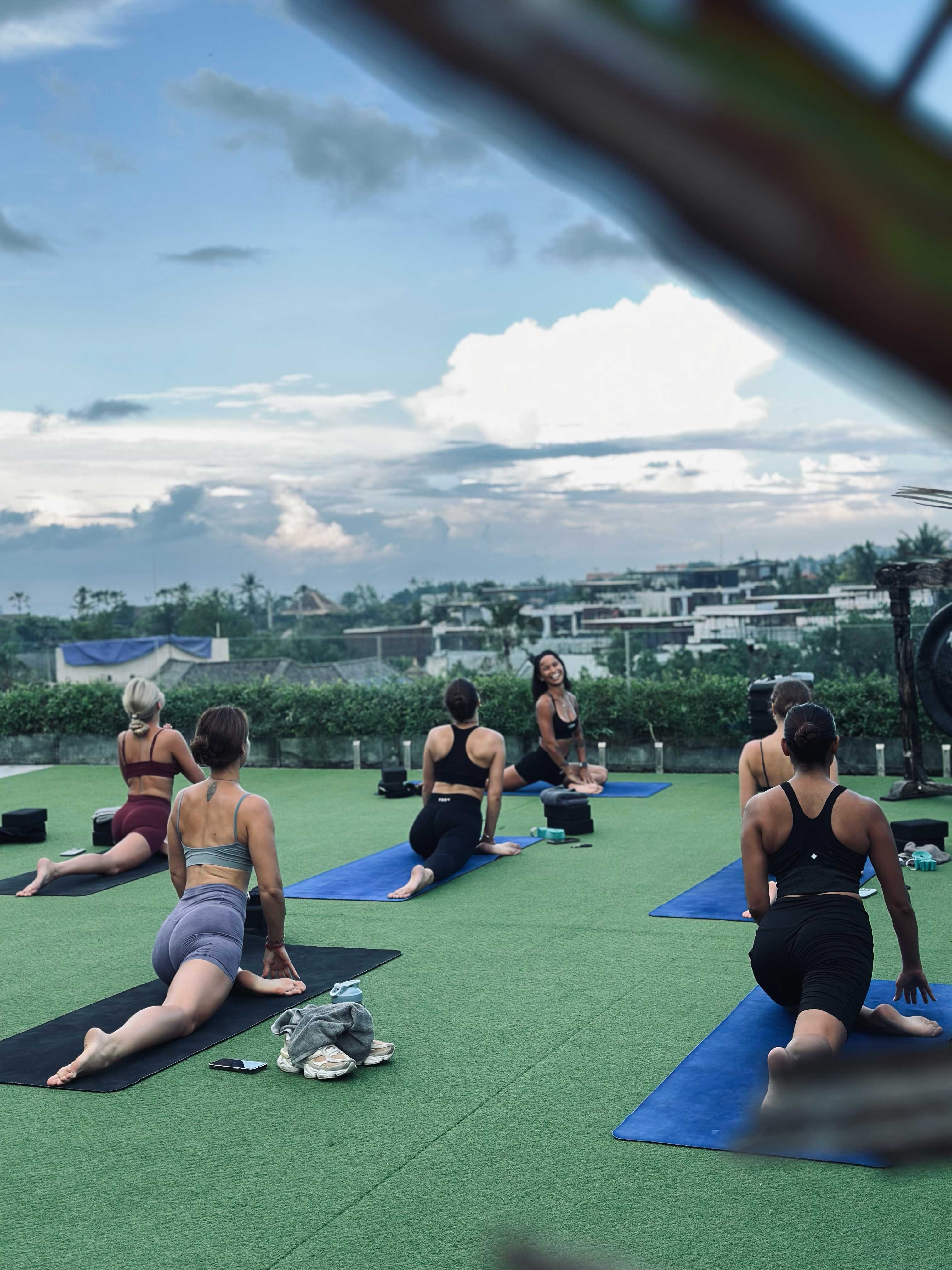 Yoga instructor smiling as a group of people practice pigeon pose on a rooftop with green turf, overlooking a scenic tropical town and dramatic cloudy sky.