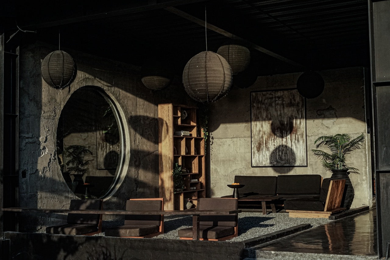 Moody, minimalist lounge area with low seating, wooden bookshelf, concrete walls, hanging lanterns, and a large round window reflecting soft afternoon light.