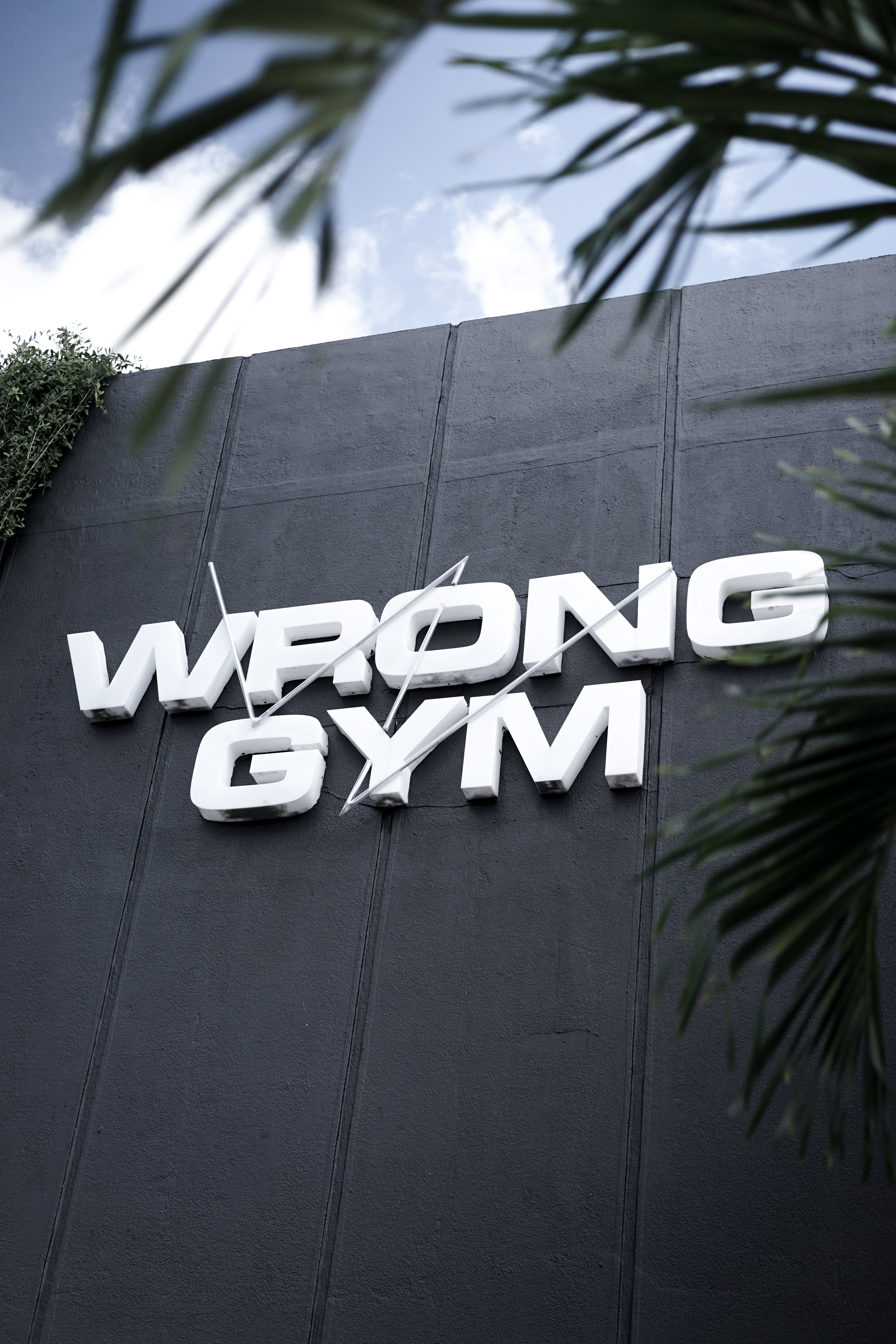 Angled view of the exterior of Wrong Gym Bali, showing a bold white 3D logo on a dark facade with hanging green vines, set against a clear blue sky.