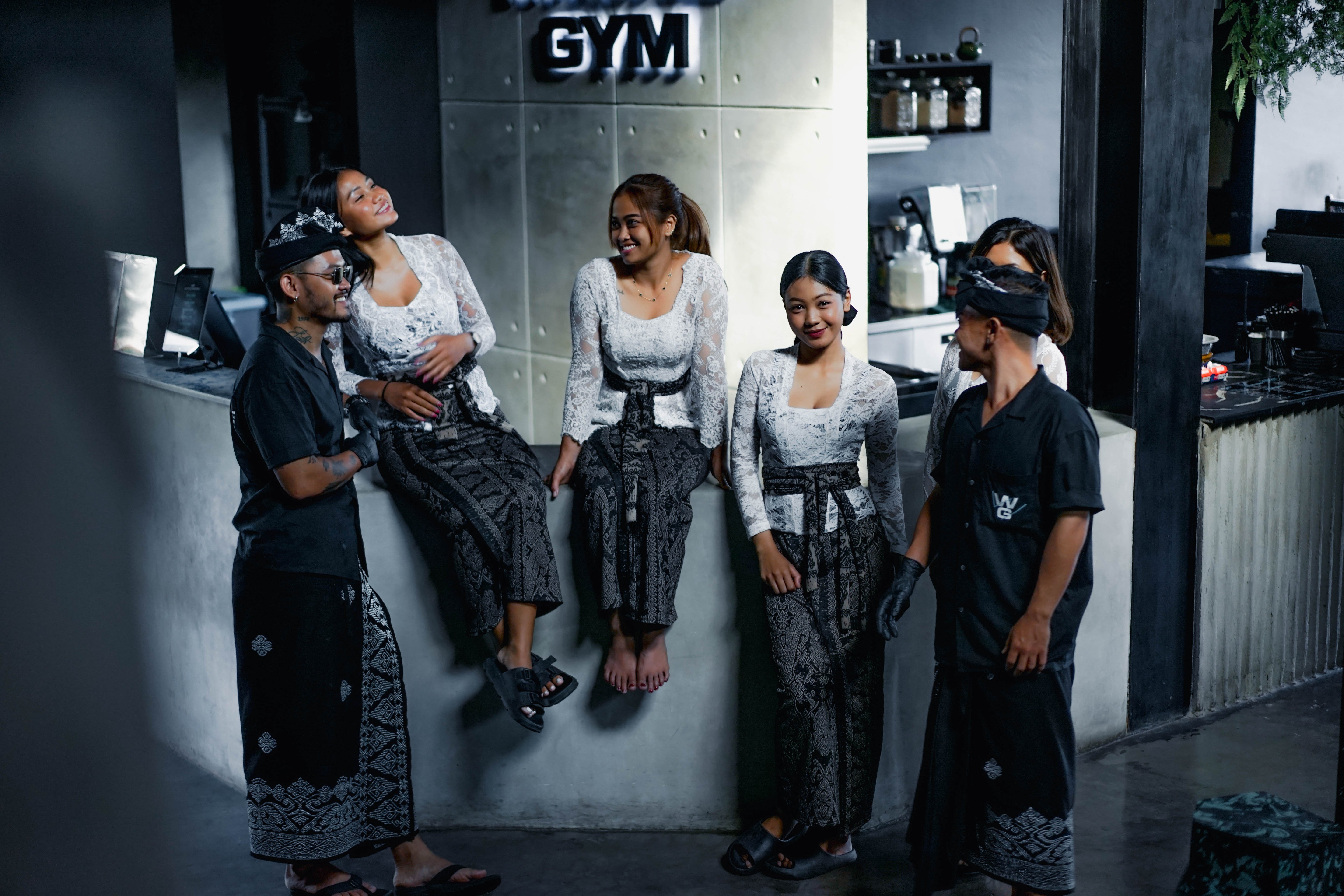 Team members at Wrong Gym wearing traditional Balinese attire, smiling and chatting near the reception counter in a modern concrete interior.