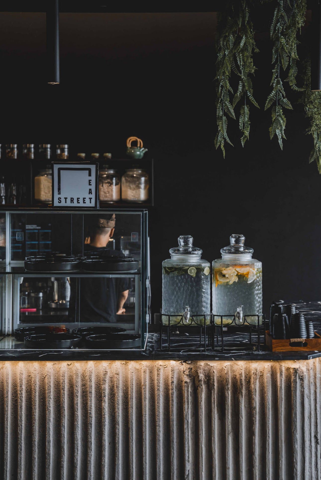Minimalist café area at Wrong Gym Bali featuring a concrete-textured counter, infused water dispensers, and a glass food shelf labeled 'Eat Street' with black plates and jars displayed in the background.
