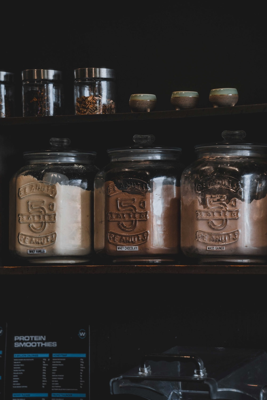Glass jars of protein powders and supplements labelled Whey Vanilla, Whey Chocolate, and Mass Gainer at the Wrong Gym Bali café counter.