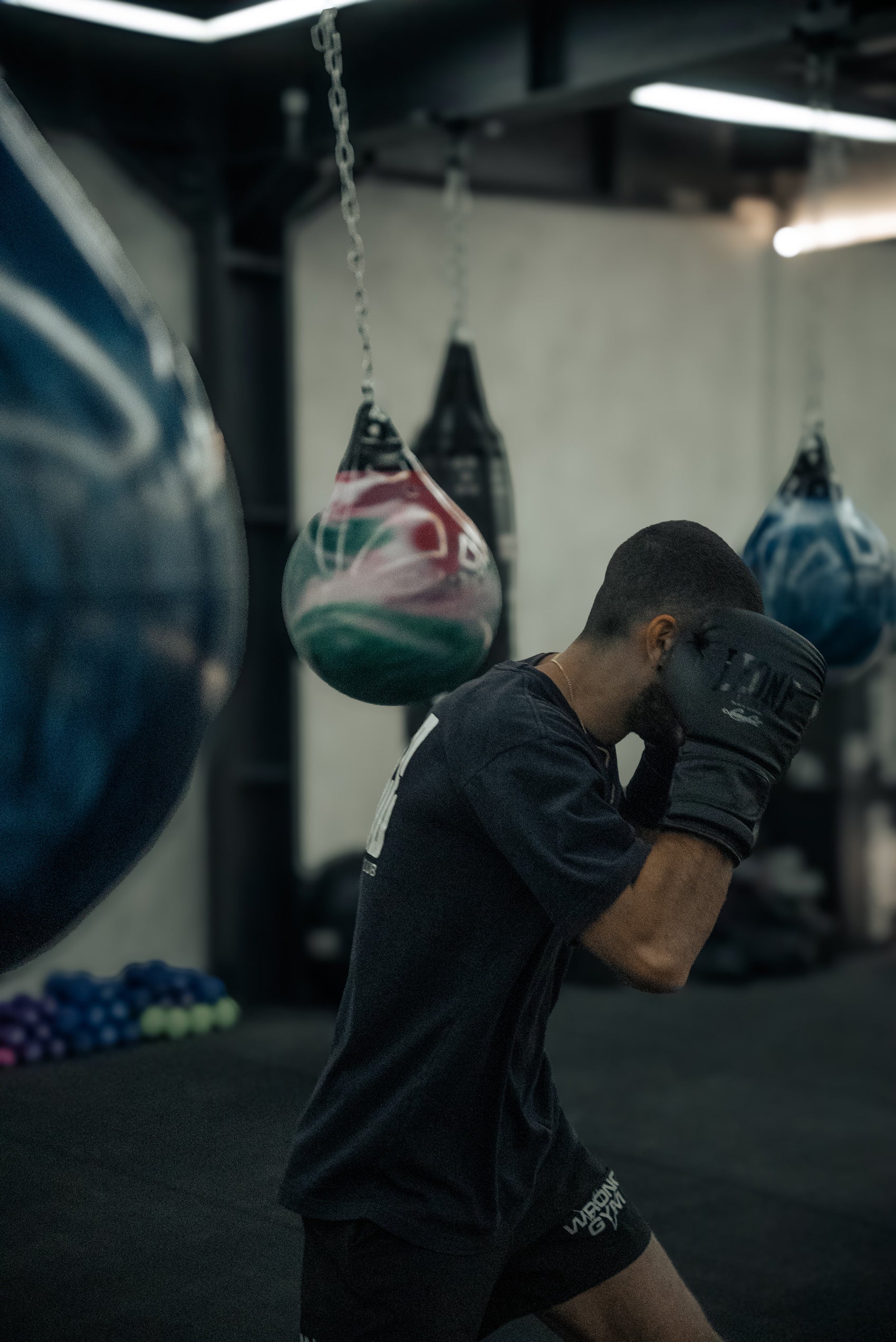 Man wearing boxing gloves and Wrong Gym Bali athletic gear training with suspended aqua punching bags in a dimly lit boxing class studio.