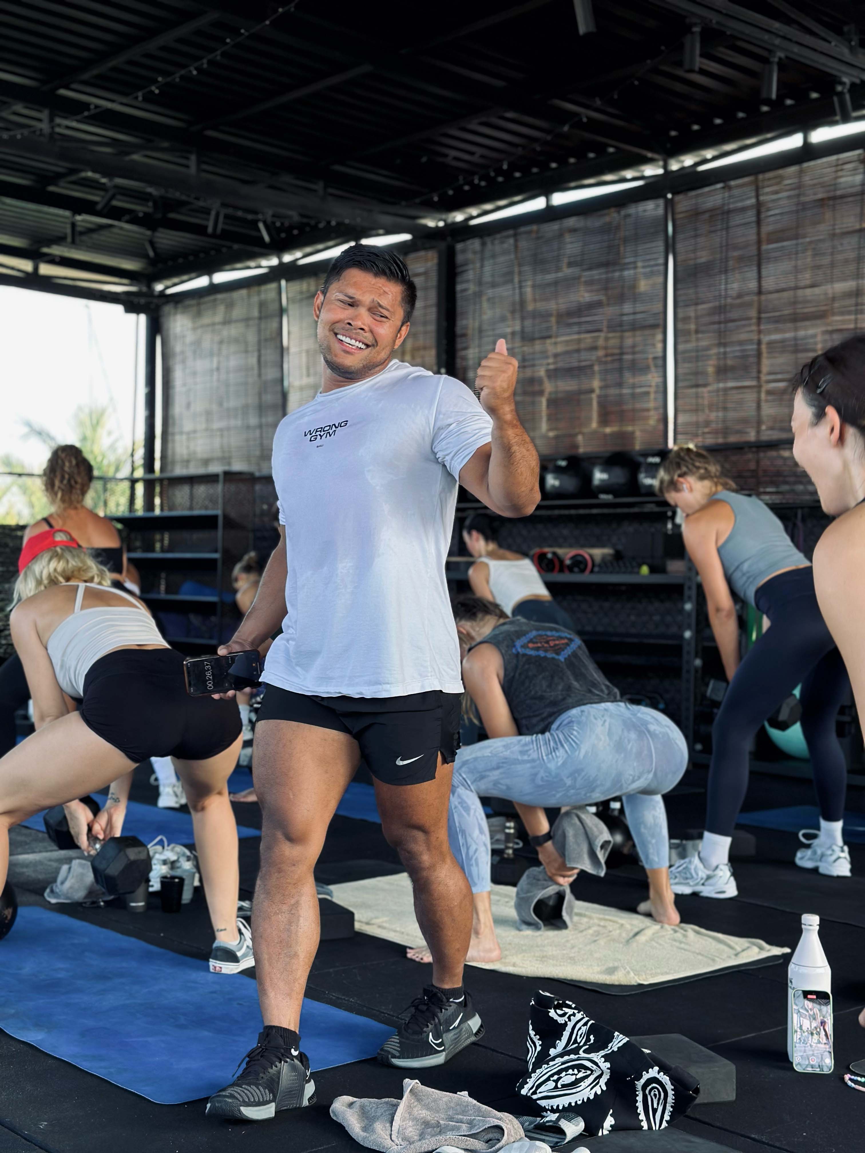 Personal trainer smiling and holding a dumbbell during a Booty Build class at Wrong Gym Bali, with a group of participants performing squats and lunges in the background.
