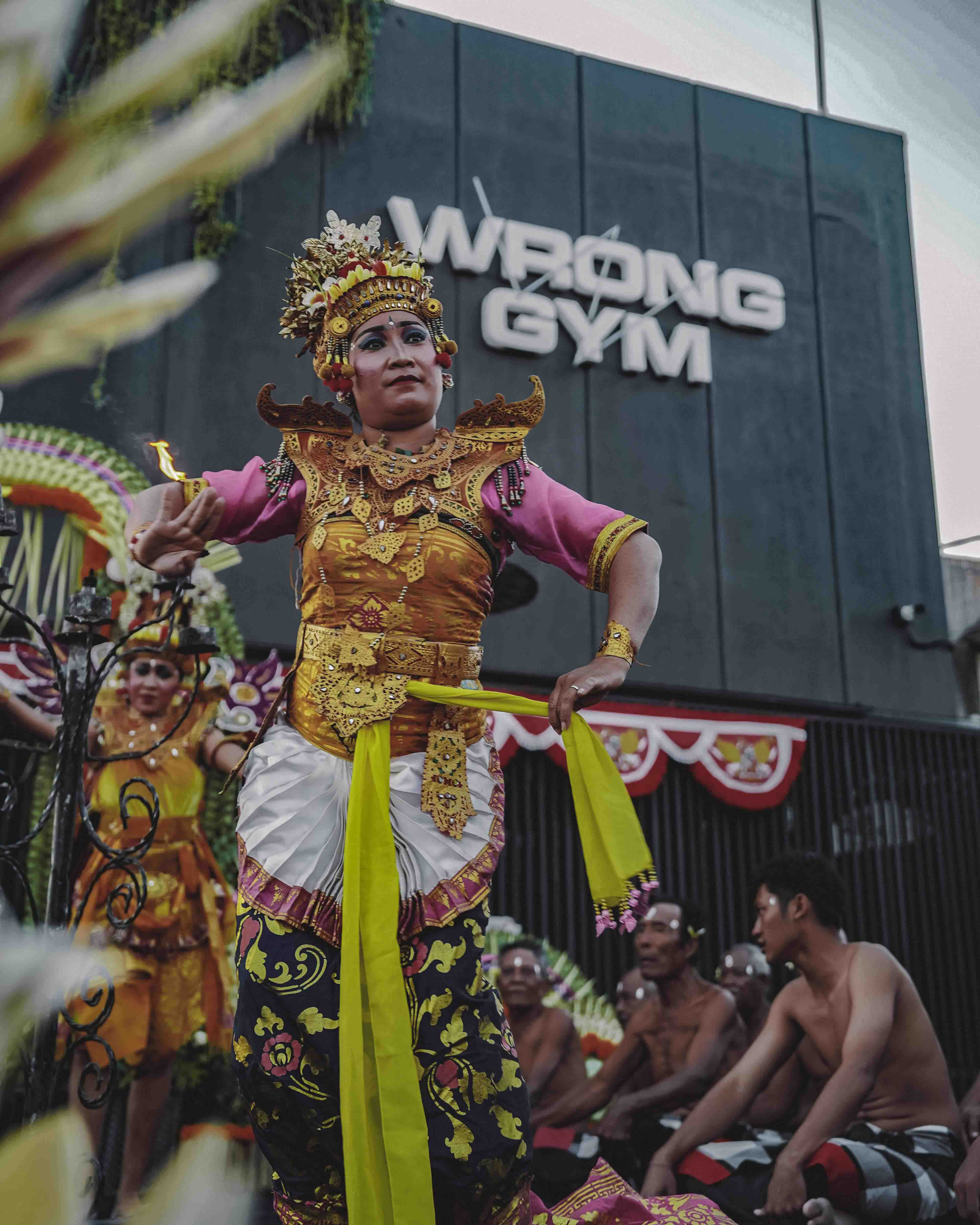 Bali dancer in traditional costume performing in front of Wrong Gym Bali during the gym's anniversary celebration, with musicians seated behind.