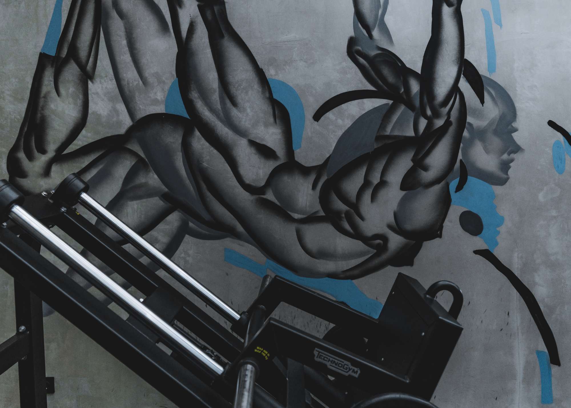 Close-up of a Technogym leg press machine in front of a dynamic mural of a muscular human figure in motion at Wrong Gym Bali.