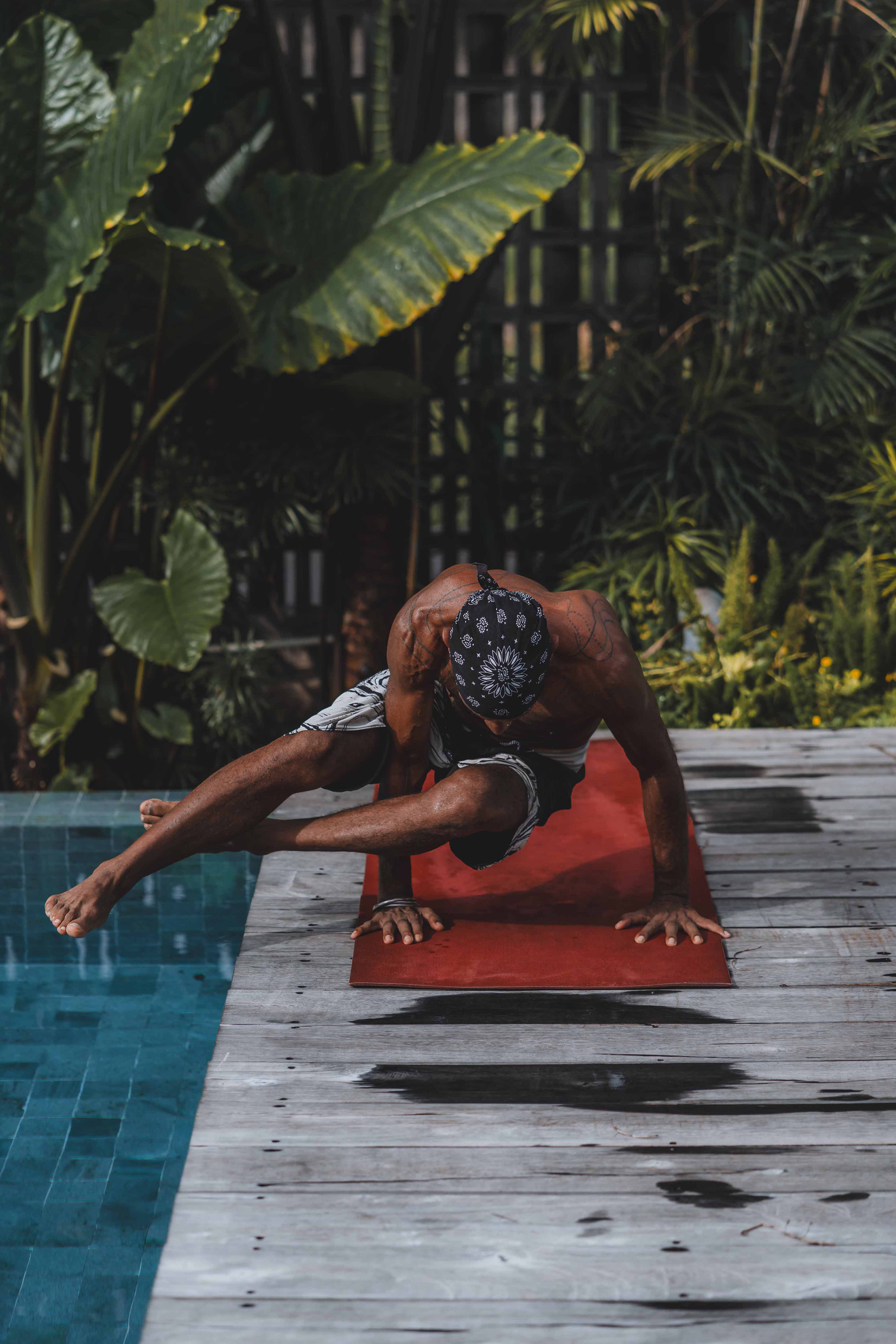 Man practising advanced yoga pose on a red mat by the poolside at Wrong Gym Bali, surrounded by tropical greenery.