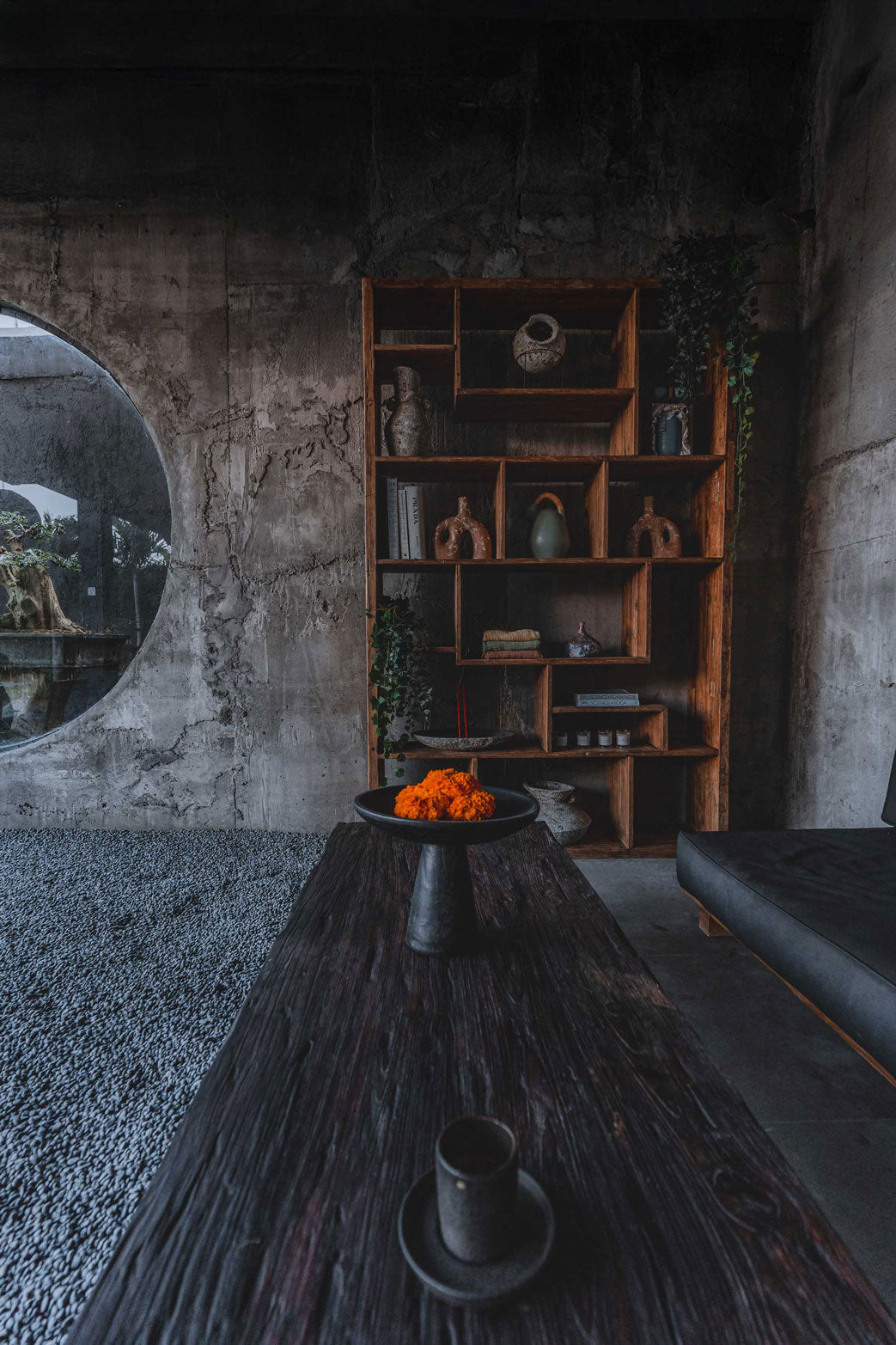 Earth-toned recovery lounge at Wrong Gym Bali featuring a rustic wooden shelf with pottery, books, and plants, next to a dark timber table with marigold flowers and ceramic cup decor.