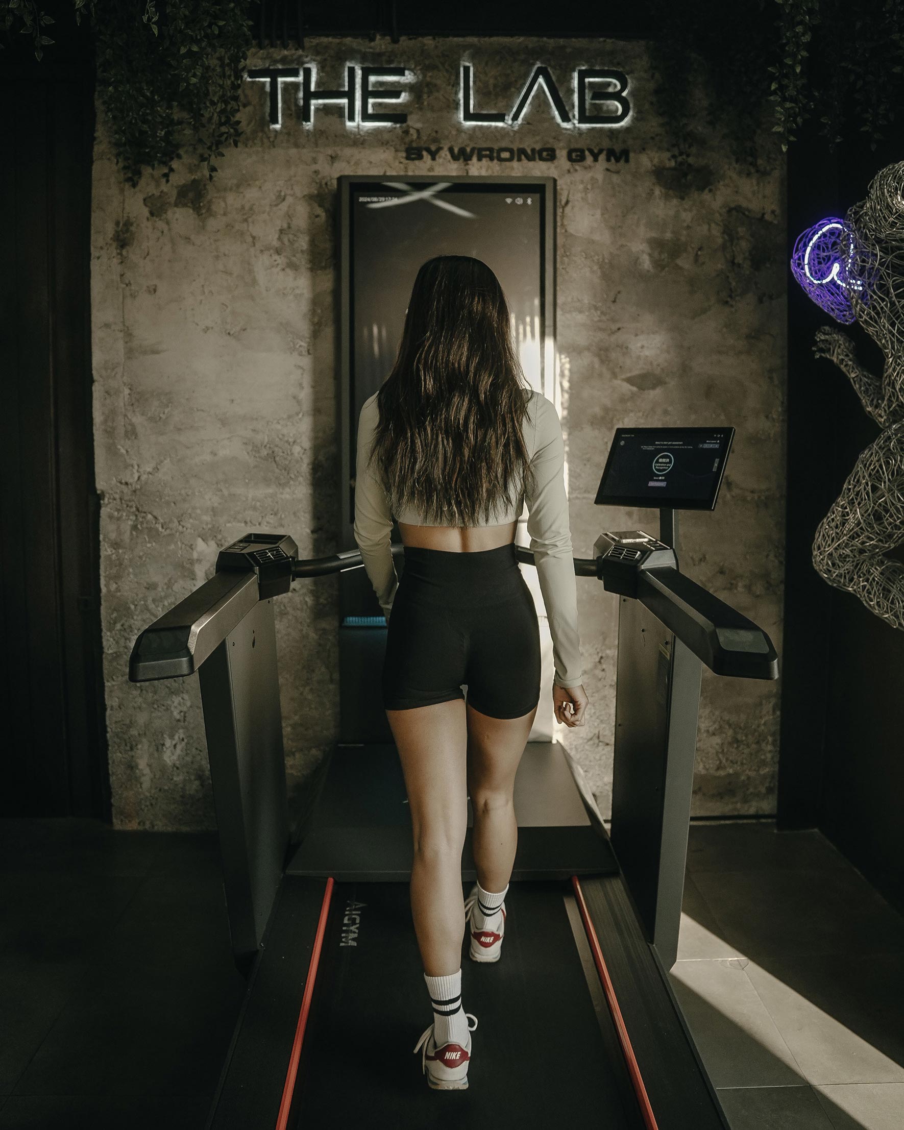Female fitness enthusiast walking on a treadmill at The Lab by Wrong Gym Bali, with neon signage and modern interior decor in view.