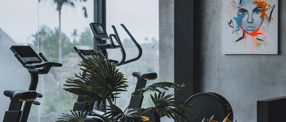 A sleek indoor gym space with stationary exercise bikes lined up in front of a large window overlooking tropical greenery. A modern art portrait is mounted on the grey concrete wall, and vibrant indoor plants add a fresh, natural touch to the scene.
