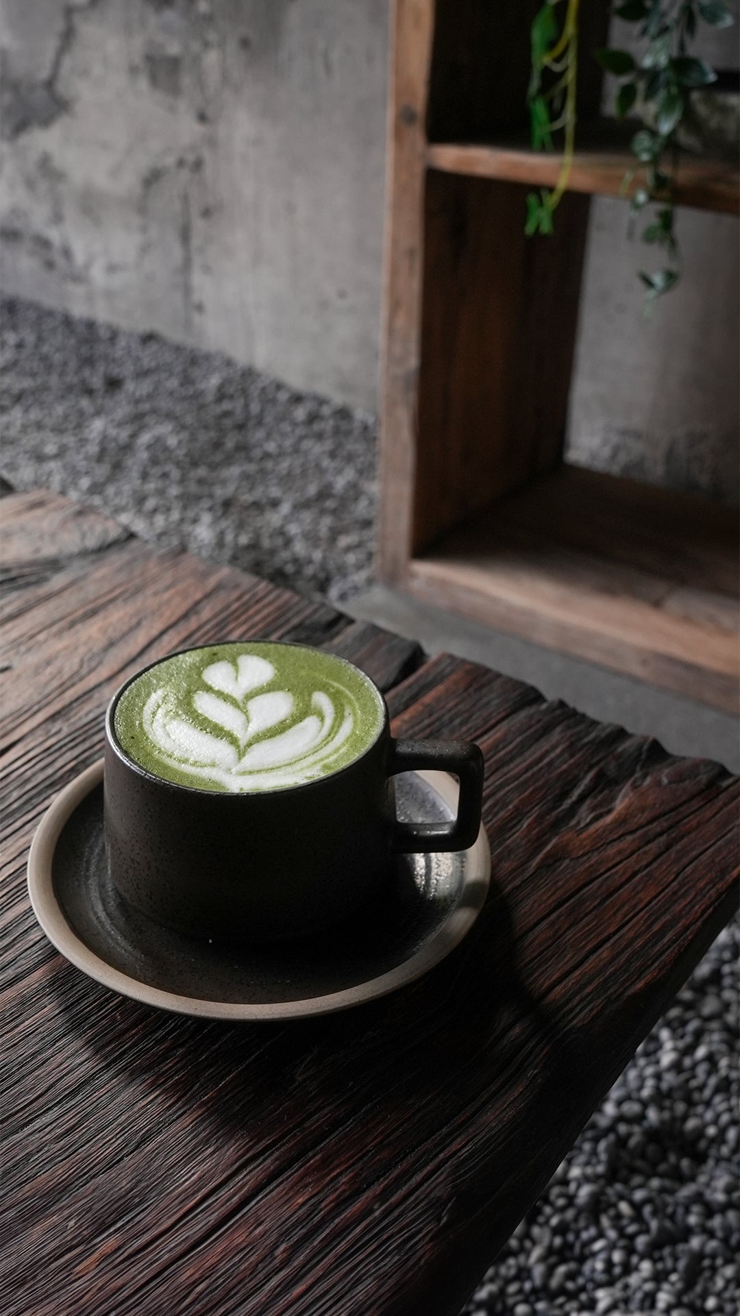"Matcha latte with heart-shaped latte art served in a black ceramic cup on a rustic wooden table, set against a stone-textured wall and minimalist decor at a modern Bali café."