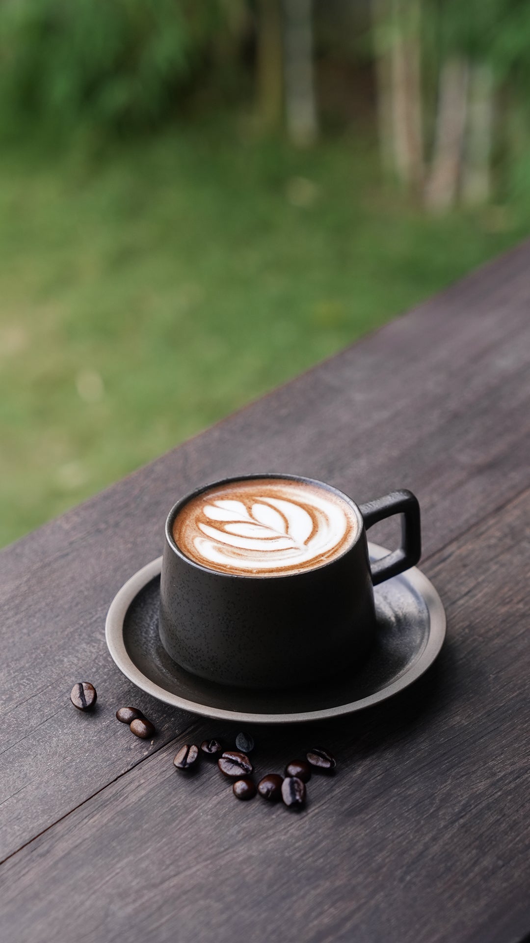 "Artisan cappuccino in a matte black ceramic cup on a matching saucer, with latte art on top and scattered coffee beans, placed on a dark wooden table overlooking a garden."