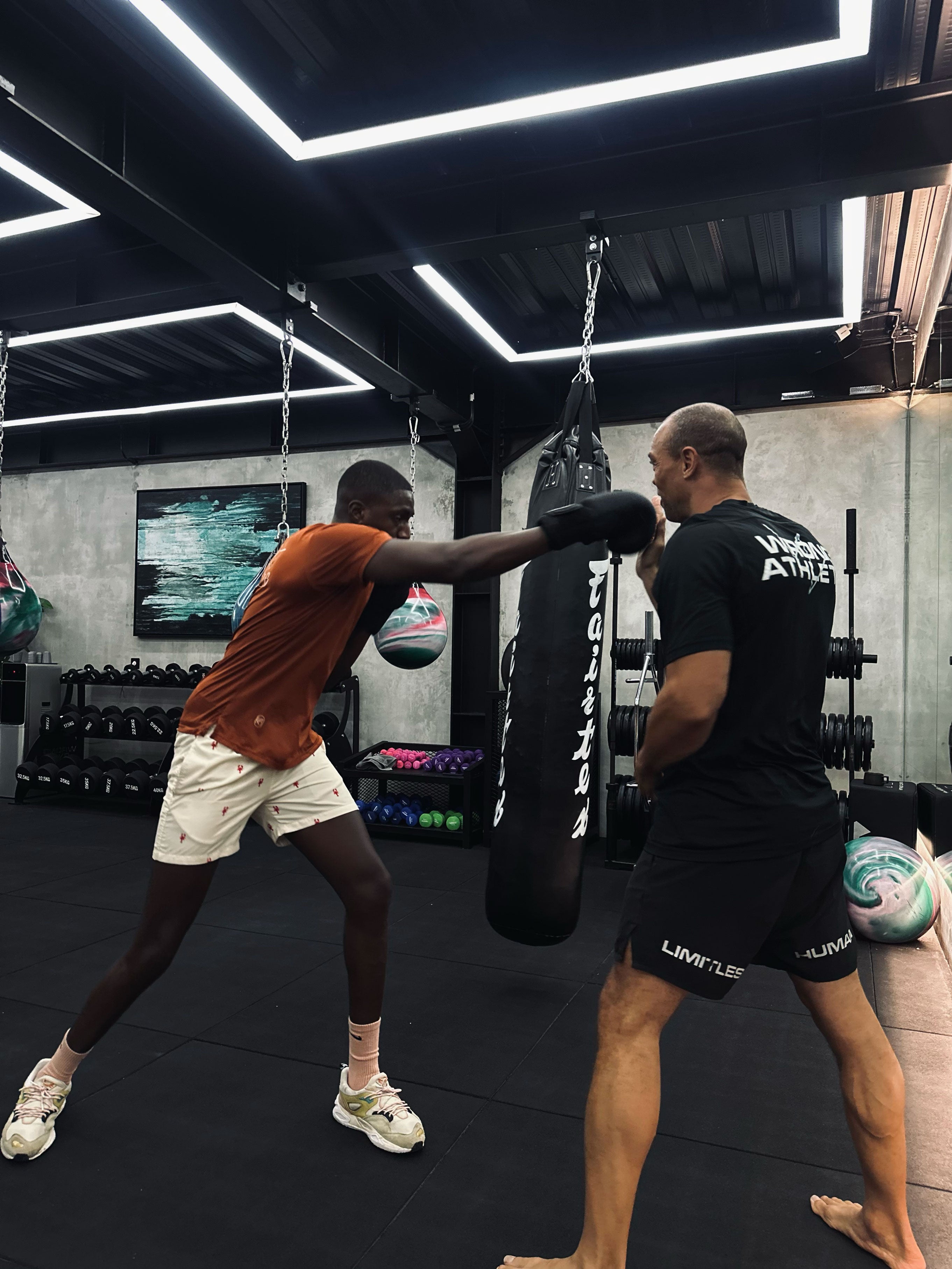 Young male athlete throwing a punch during a boxing training session with a coach inside a modern, neon-lit gym at Wrong Gym Bali, surrounded by hanging aqua bags and free weights.