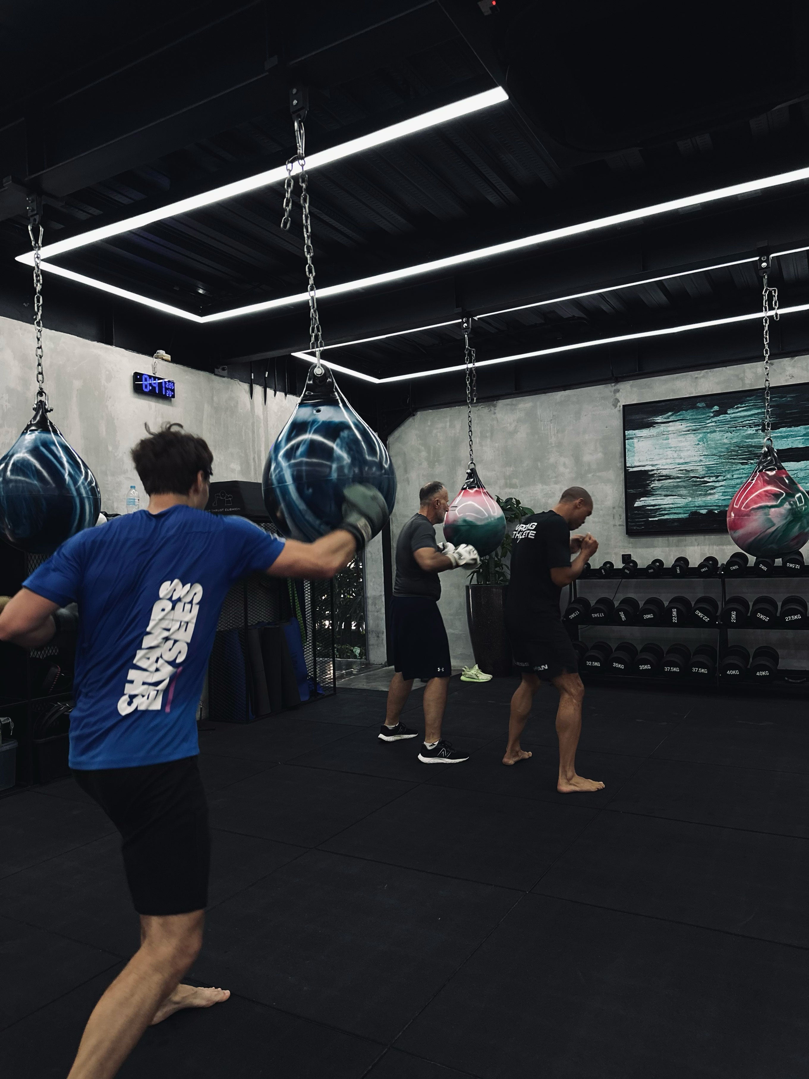 Group of men engaged in a boxing workout session at Wrong Gym Bali, practicing strikes with aqua punching bags in a sleek, dimly lit training room equipped with free weights and padded flooring.