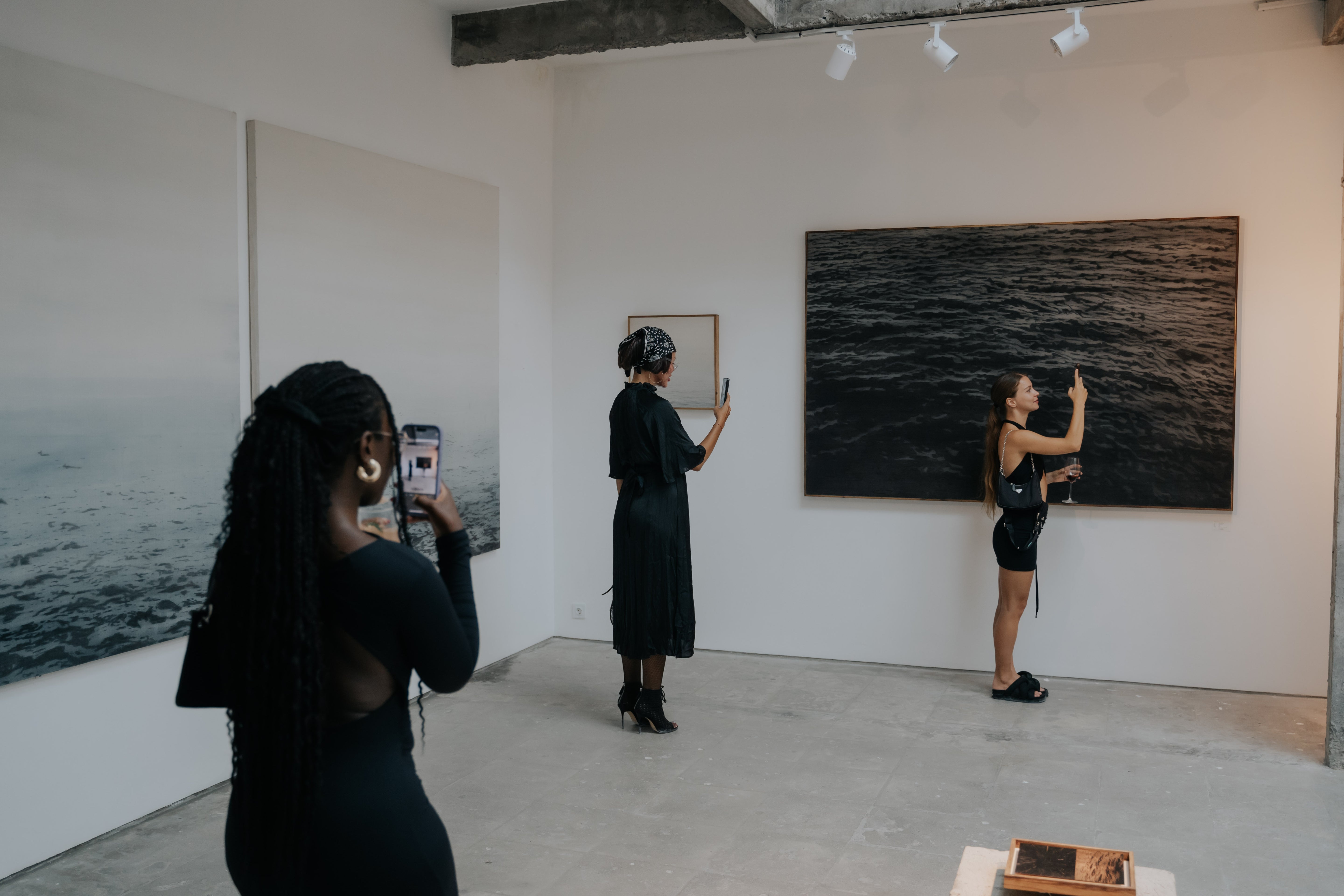 Three women in stylish black outfits explore a minimalist art gallery featuring ocean-themed paintings. Each woman is capturing artwork on their phone, with one holding a glass of wine. The gallery has a clean, industrial interior with concrete floors and white walls, evoking a modern, curated ambiance.