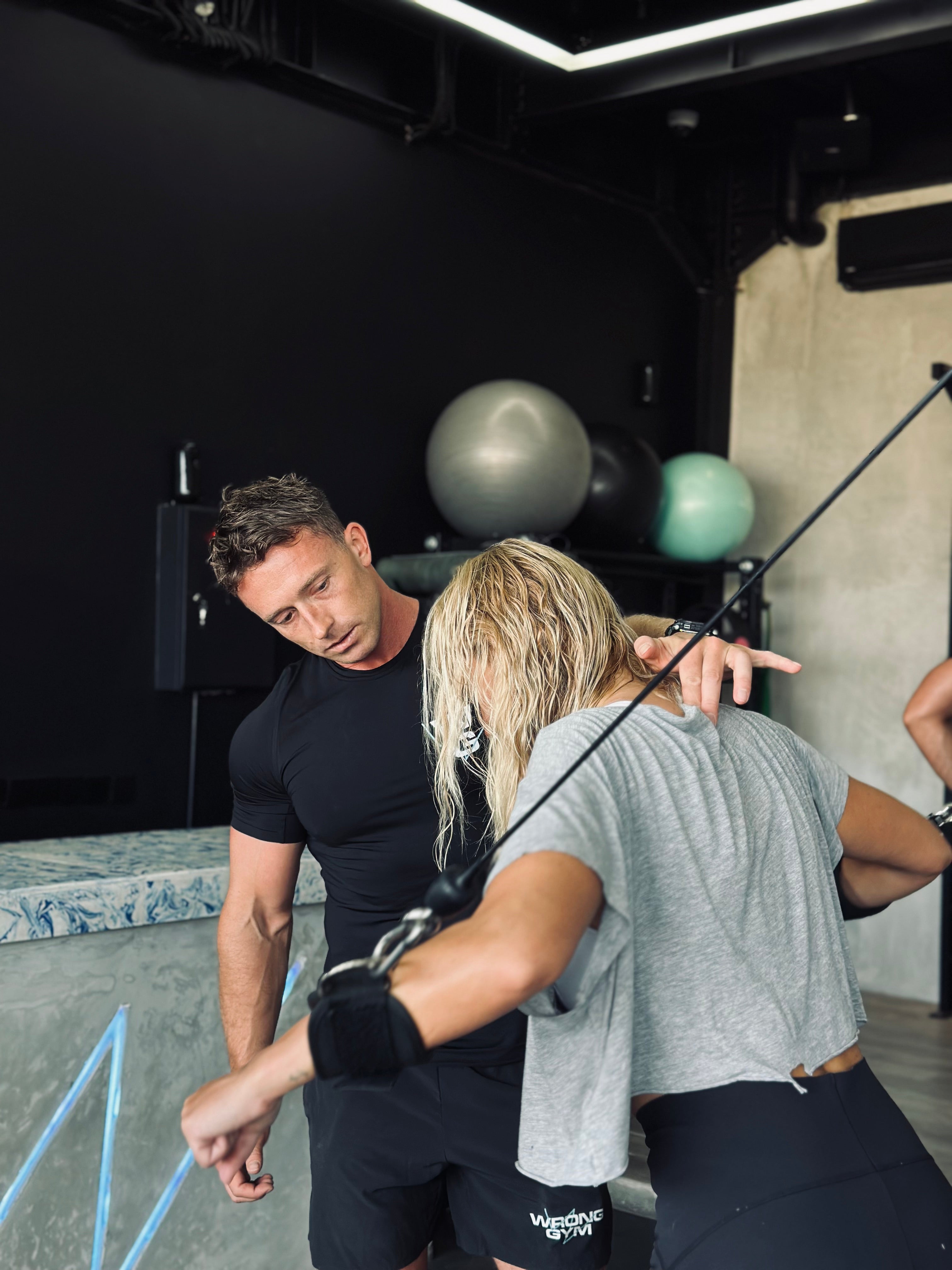 A personal trainer at Wrong Gym Bali closely guides a woman during a resistance cable workout. The trainer observes her form attentively, ensuring proper technique. The gym setting features a sleek, modern interior with black walls, fitness balls in the background, and a marble-style counter.