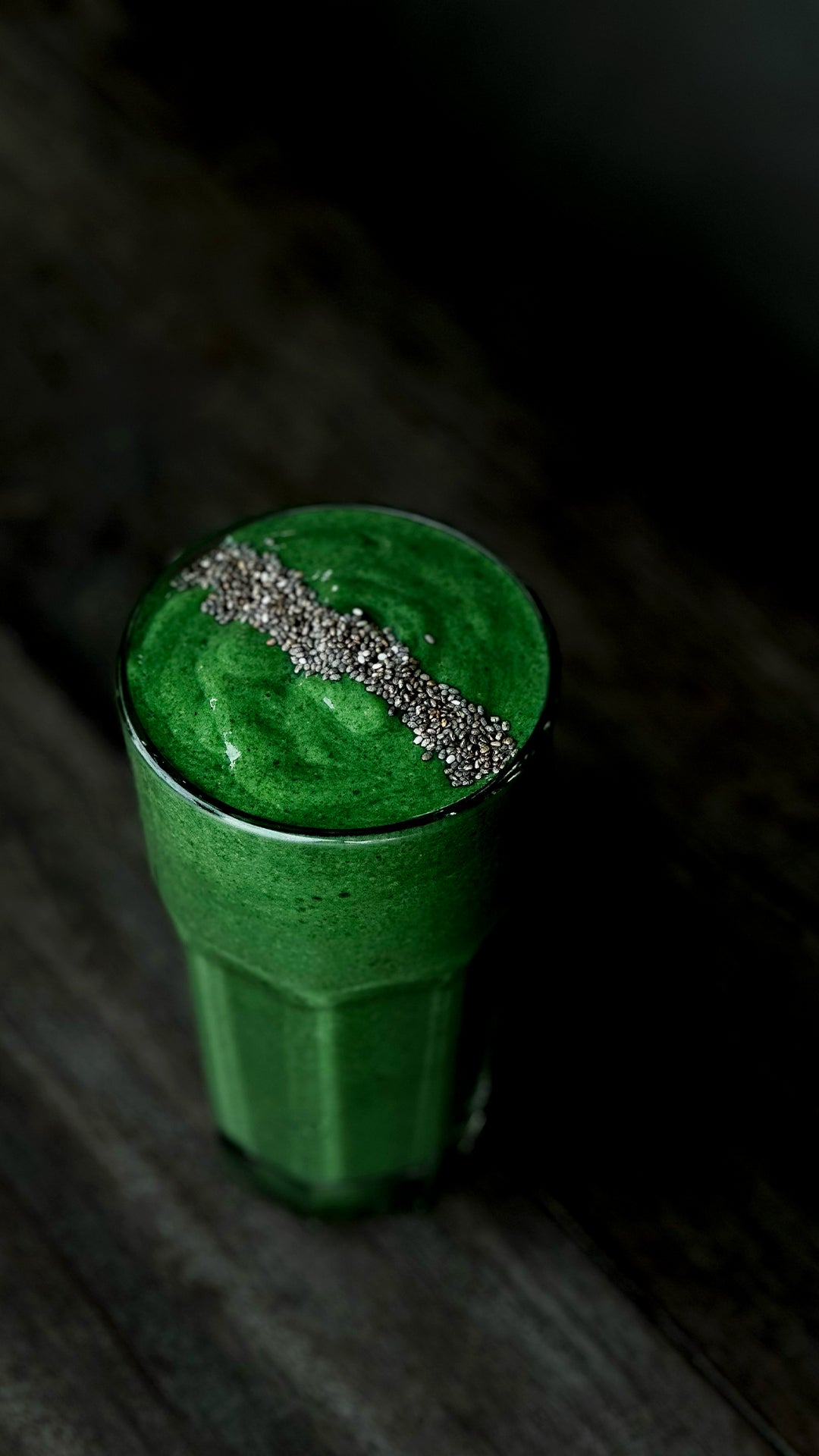 A tall glass of vibrant green protein smoothie, topped with a neat row of chia seeds, set against a dark wooden background.