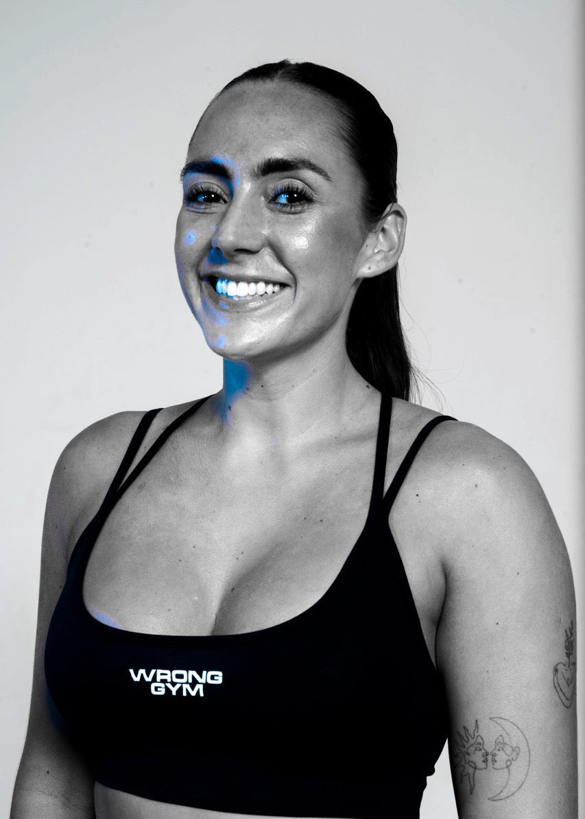 Hannah, trainer at Wrong Gym Bali, smiling confidently in a branded sports bra with visible arm tattoos.