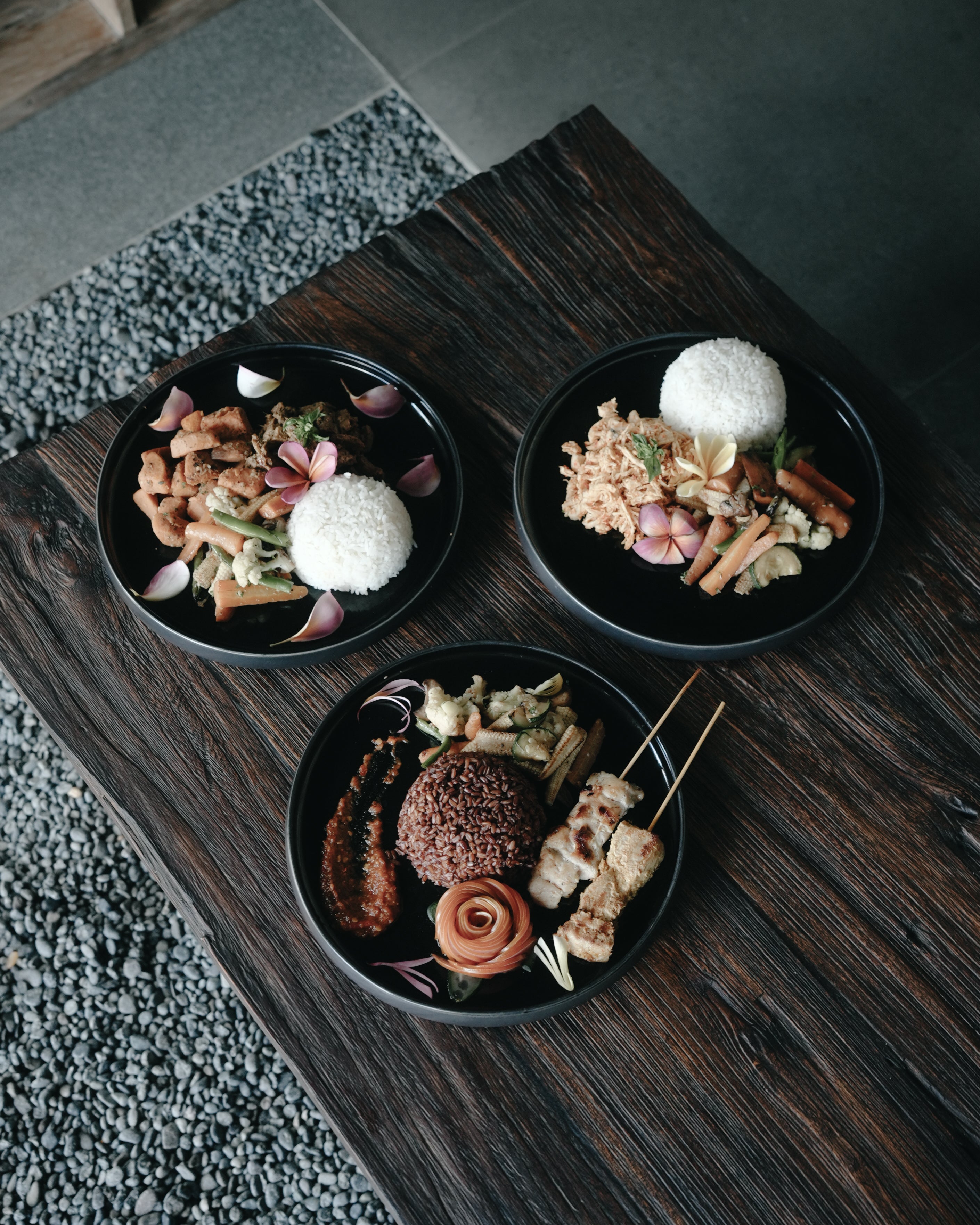 Top-down view of three black plates with Indonesian-style rice dishes, vegetables, skewers, and garnishes, served on a rustic wooden table with a stone floor background.