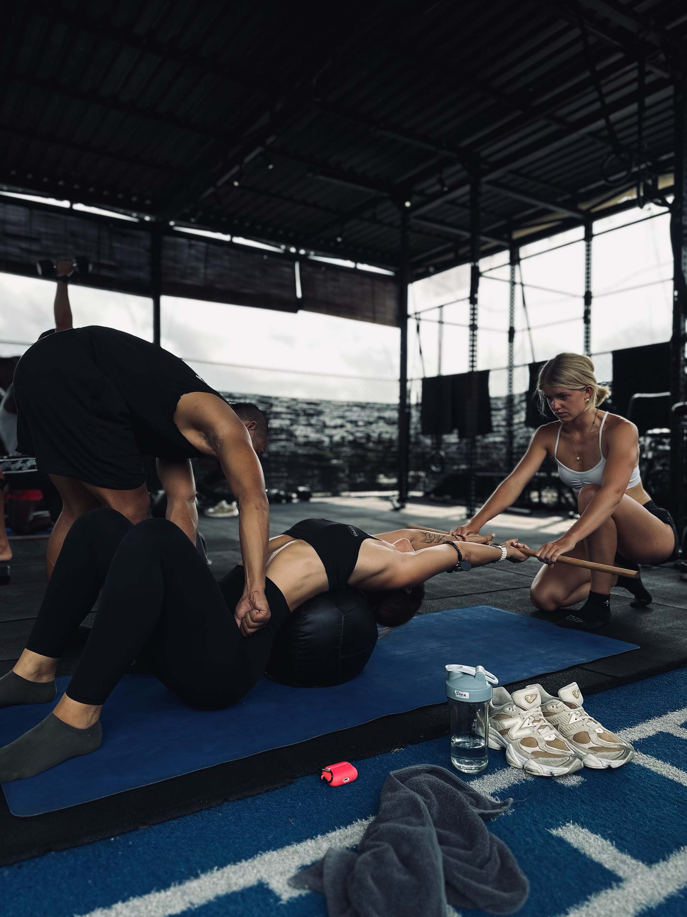 Trainers assisting a woman with a lower and upper body mobility stretch using a yoga mat and mobility tools at Wrong Gym Bali.