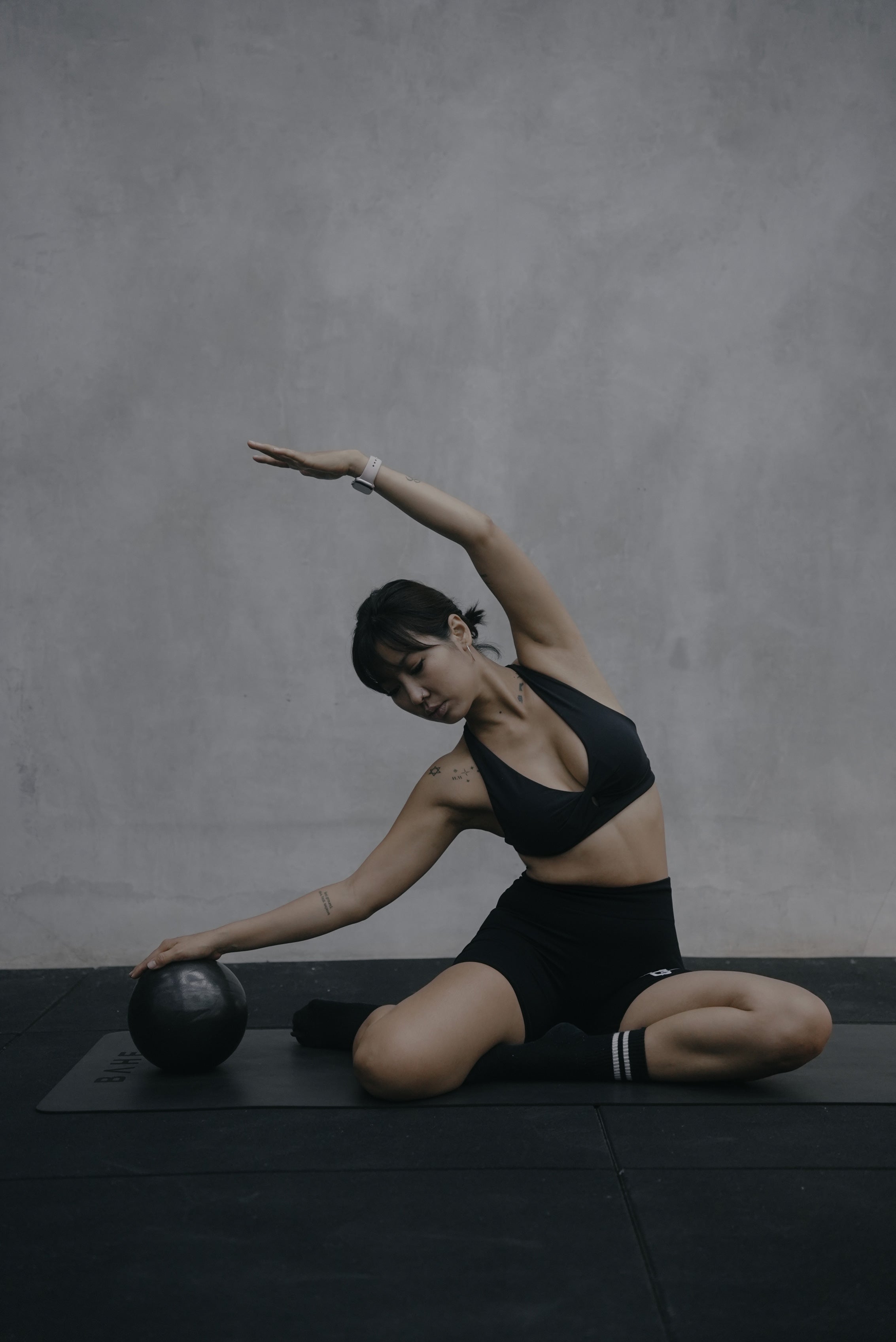 Woman in black activewear performing a seated side stretch with a medicine ball during a Pilates class at Wrong Gym Bali, set against a minimal concrete wall backdrop