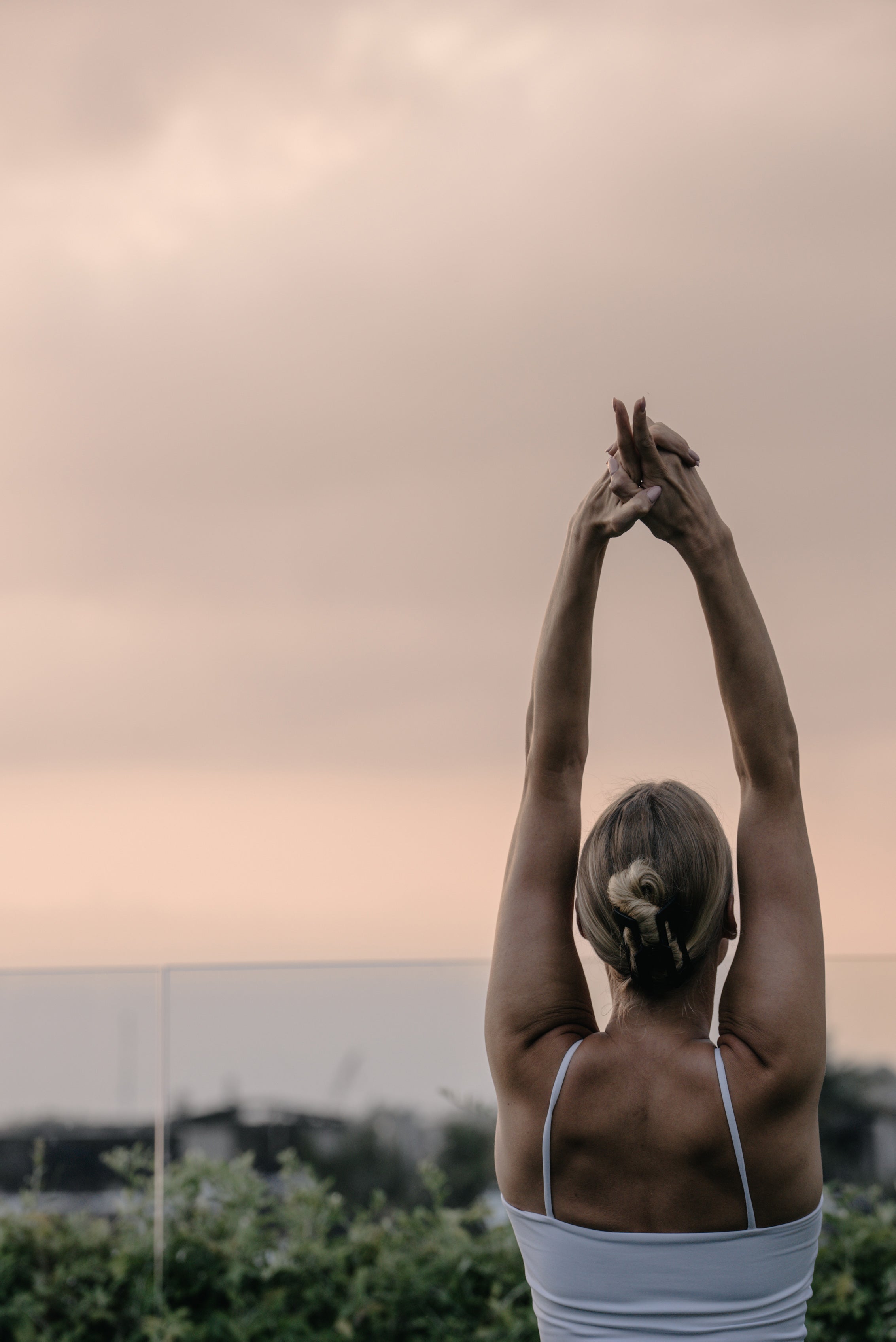 Back view of a woman in a white top stretching with arms overhead during a yoga class at sunrise on the rooftop of Wrong Gym Bali.